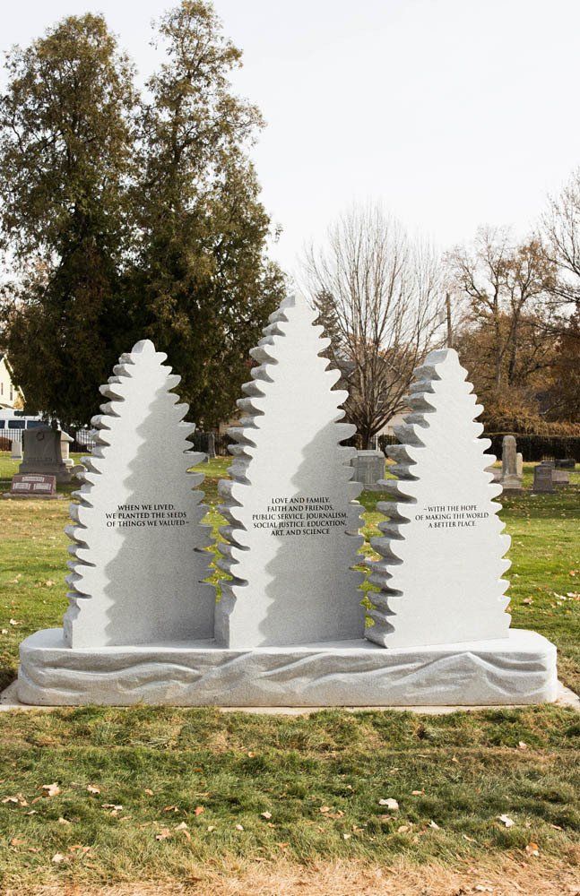 Unique pine tree-shaped white headstones aligned in a row with engraved names and a wintery background of bare trees.