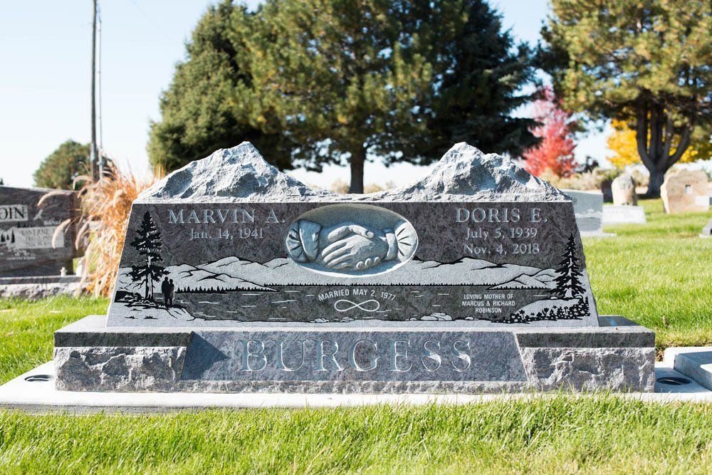 Polished black and gray granite headstone for Marvin A. and Doris E. Burgess, featuring a detailed etched landscape, a pair of clasped hands, and personalized inscriptions.