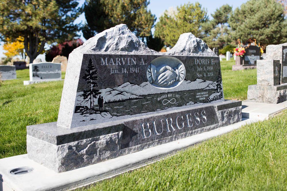 Polished black granite headstone with etched mountain scene and name “Burgess,” featuring contrasting silver engraving and a floral tribute nearby.