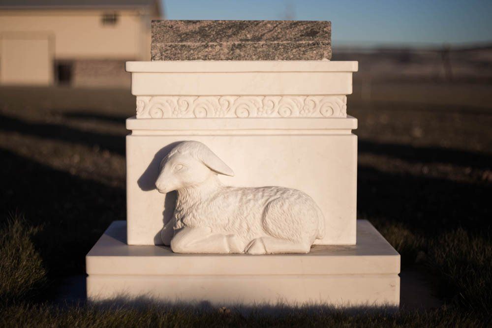 Another angle of a cream-colored stone base with a sculpted lamb, capturing a serene, pastoral cemetery view.