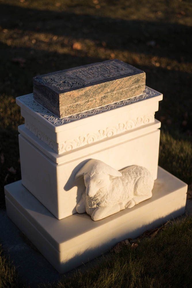Close-up of a cream-colored memorial base featuring a carved lamb, symbolizing innocence and peace, in warm afternoon light.
