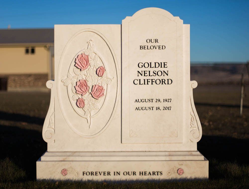Beige upright headstone with a carved oval panel of red roses and text honoring Goldie Nelson Clifford, set against a blurred cemetery background.