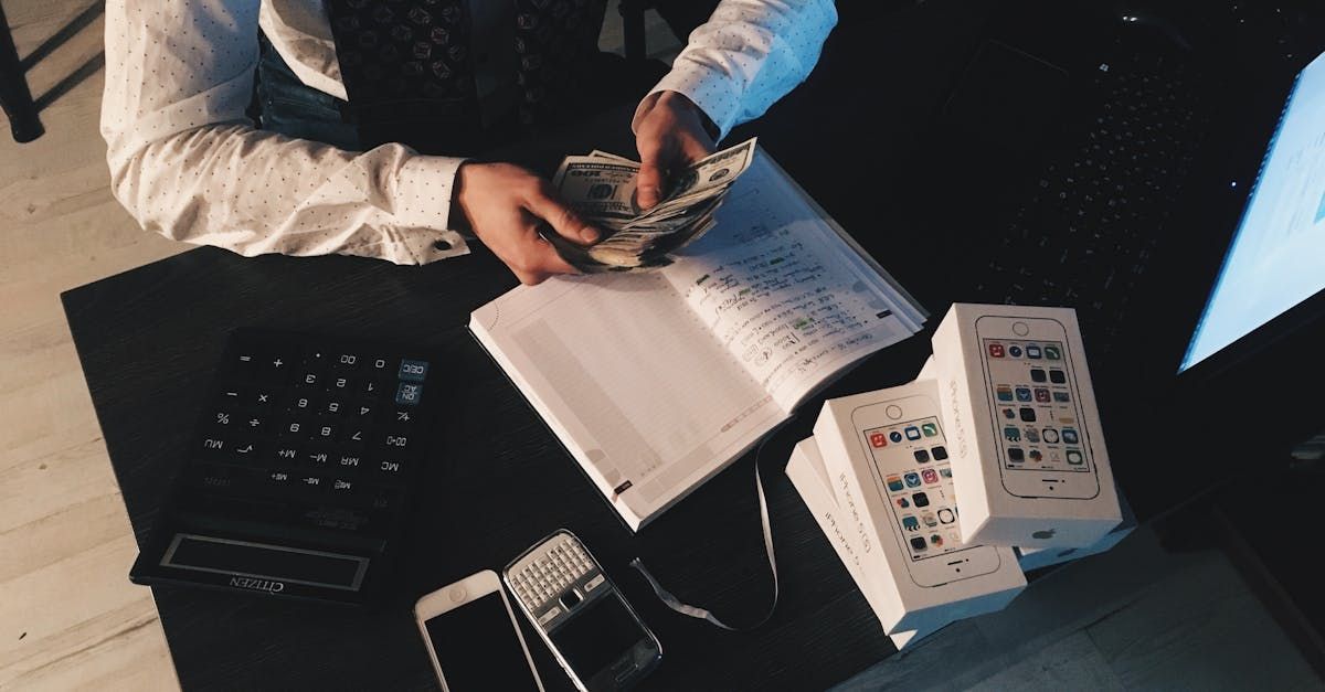 Person counting money at a desk with a notebook, phone, calculator, and stacked boxes.