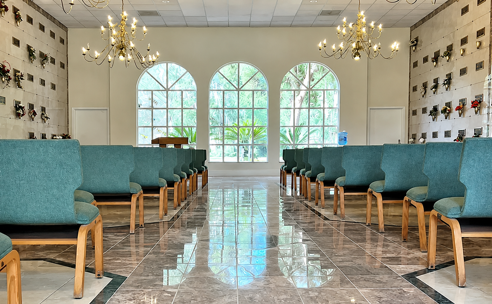Bright chapel aisle with teal chairs, chandeliers, and arched windows at the far end