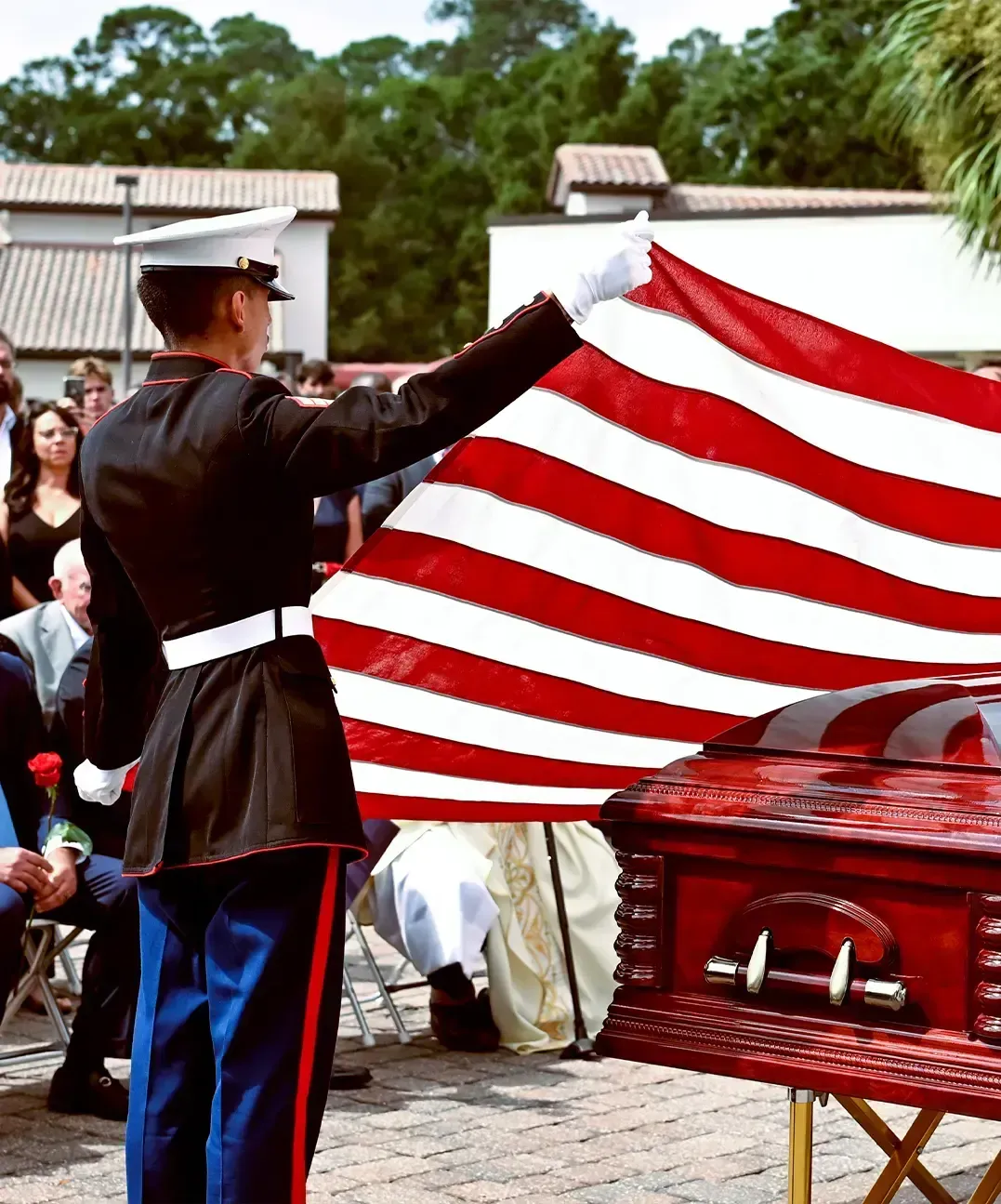 Uniformed guard unfolds a large American flag beside a red casket at an outdoor memorial ceremony