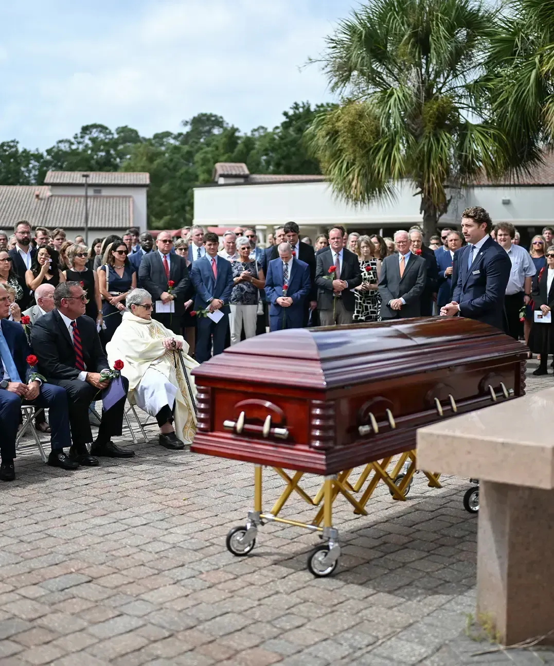 Funeral scene with a brown casket on a wheeled stand, mourners seated nearby outdoors