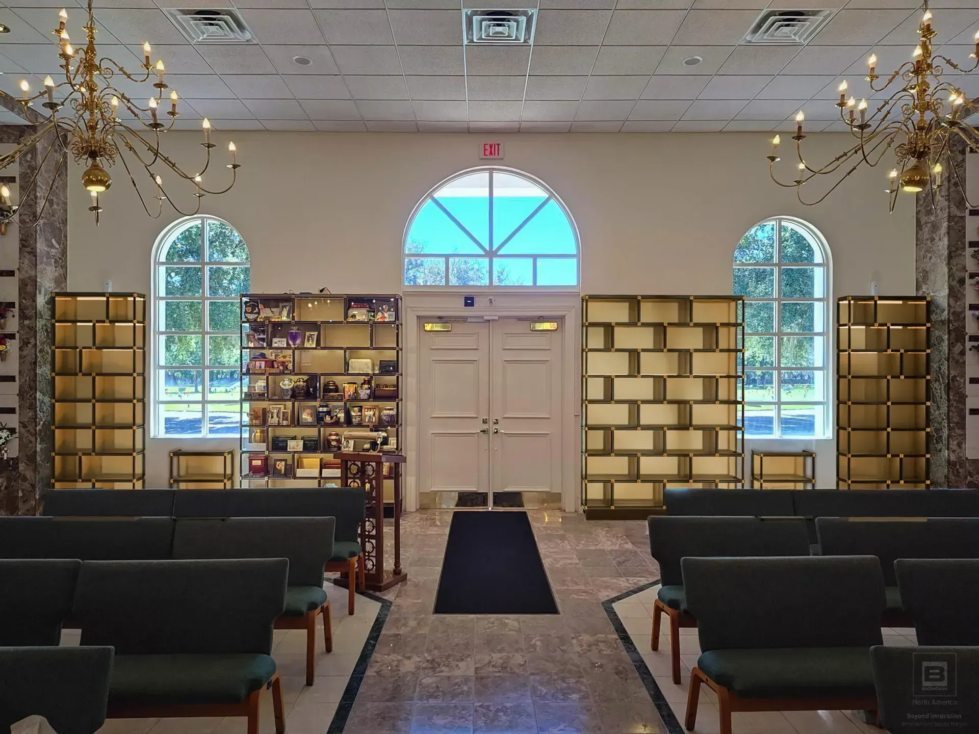 Small chapel interior with pews, a central aisle, arched windows, and a white door at the front.