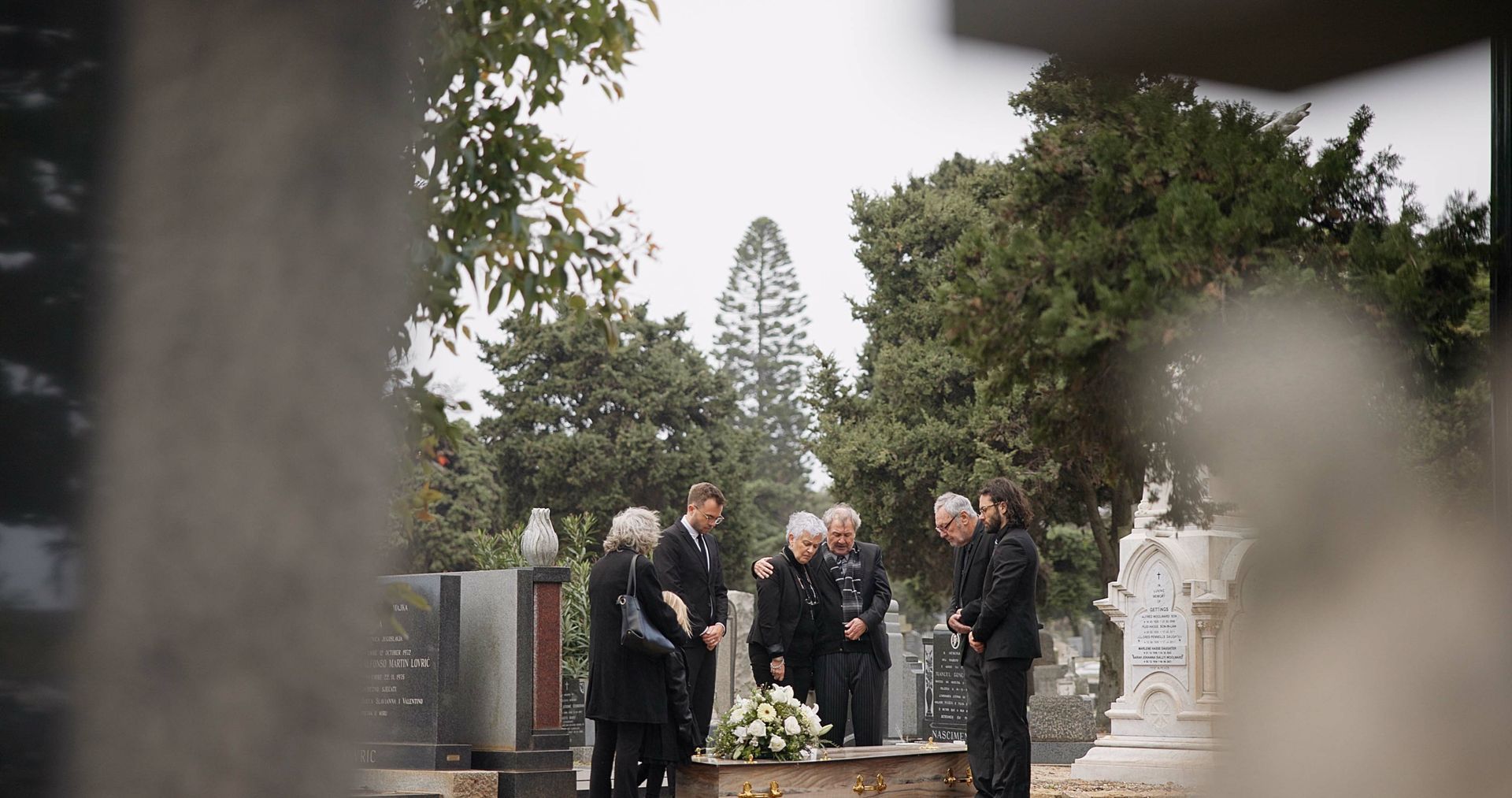Mourners gathered at a graveside ceremony in a cemetery, standing around a flower-covered casket.