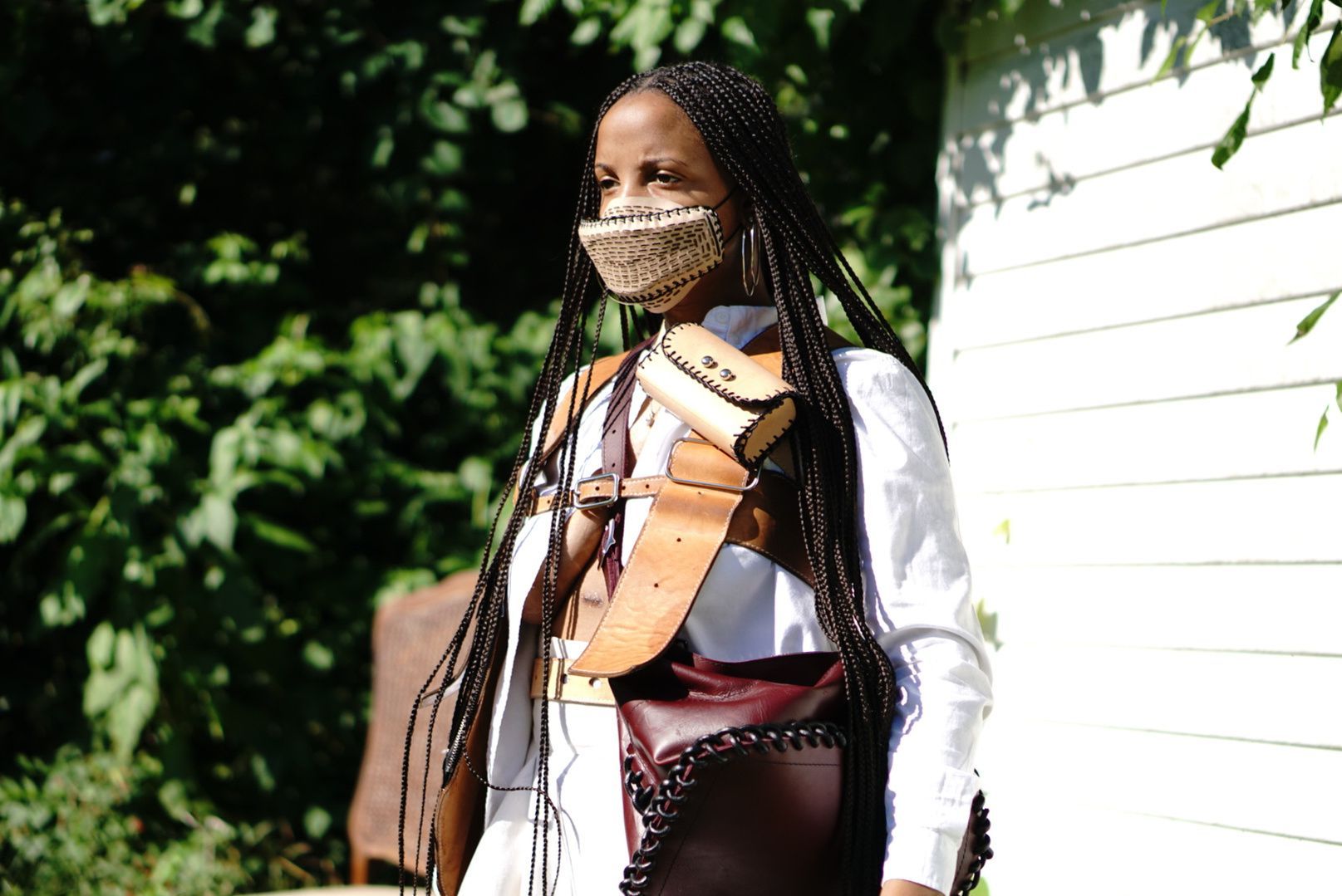 A woman wearing a mask draped in leather accessories is standing in front of a white building.