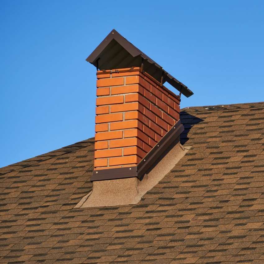 Brick chimney on a brown shingled roof against a clear blue sky.