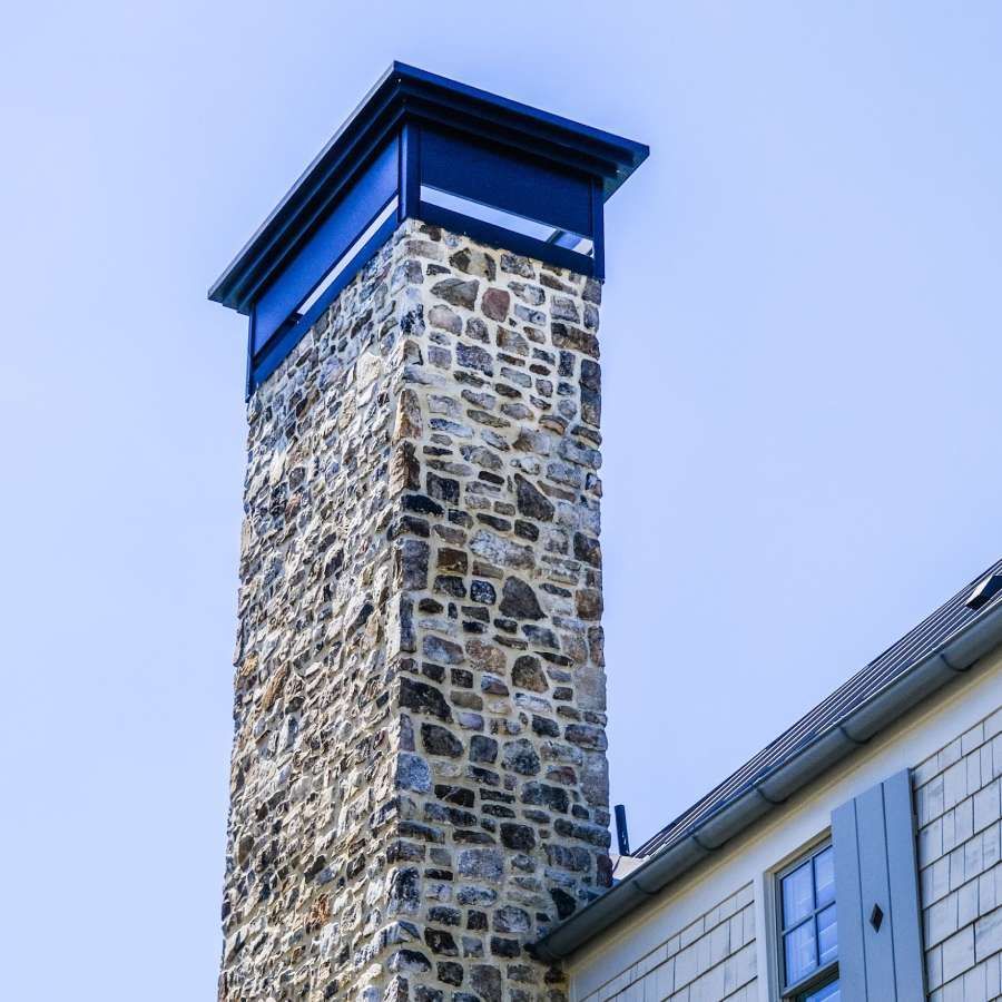 Stone chimney with dark metal cap against a clear blue sky, next to a white building.