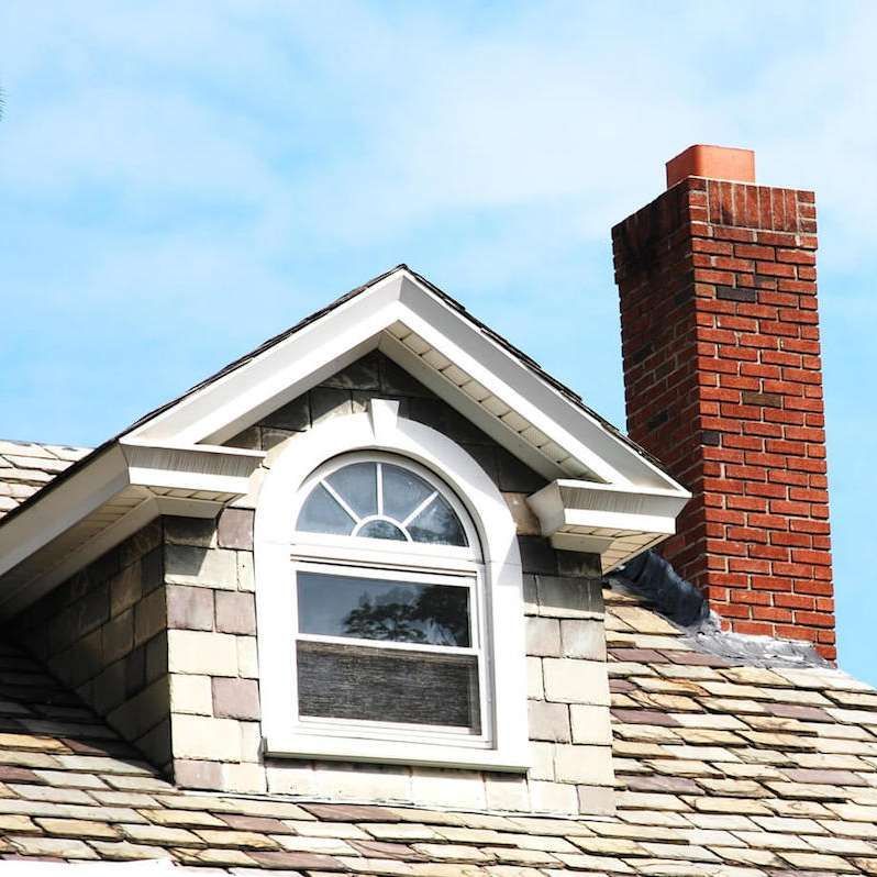 Dormer window with arched top and brick chimney on a house roof against a blue sky.