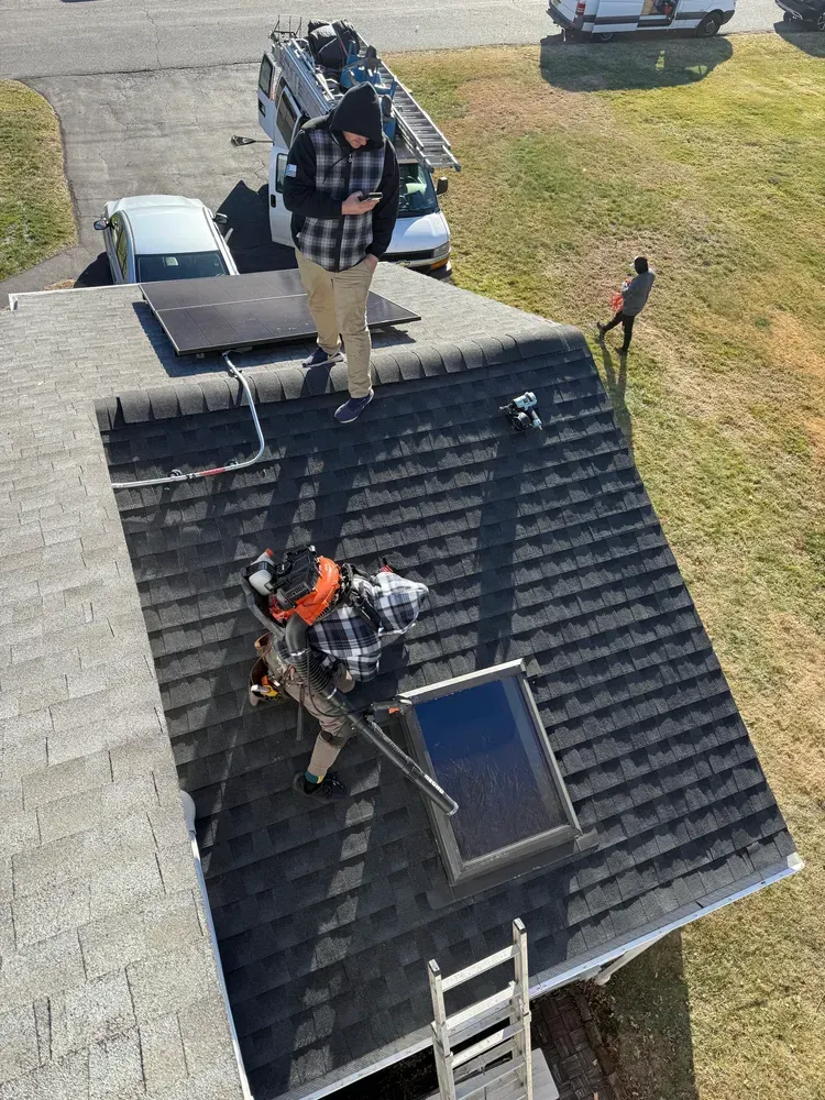 Workers installing solar panels on a roof, with a ladder and vehicle nearby.