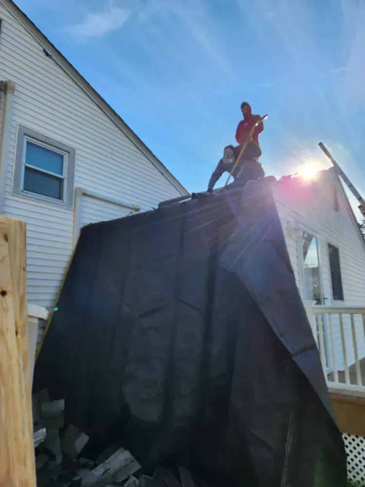 Two people on a roof covered with a black tarp, working on a house under a sunny sky.