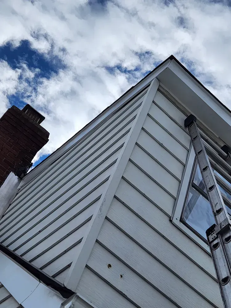 White siding on a house with a chimney, ladder, and a blue cloudy sky.