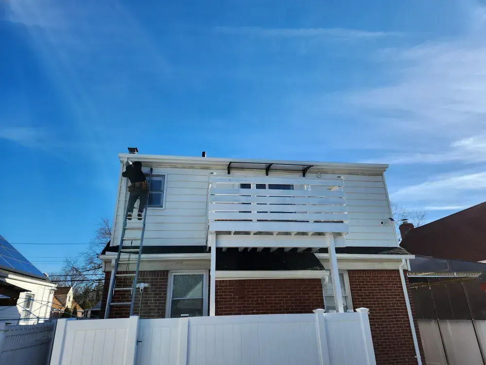 Person on ladder painting white trim on a two-story building with a deck. Blue sky in the background.