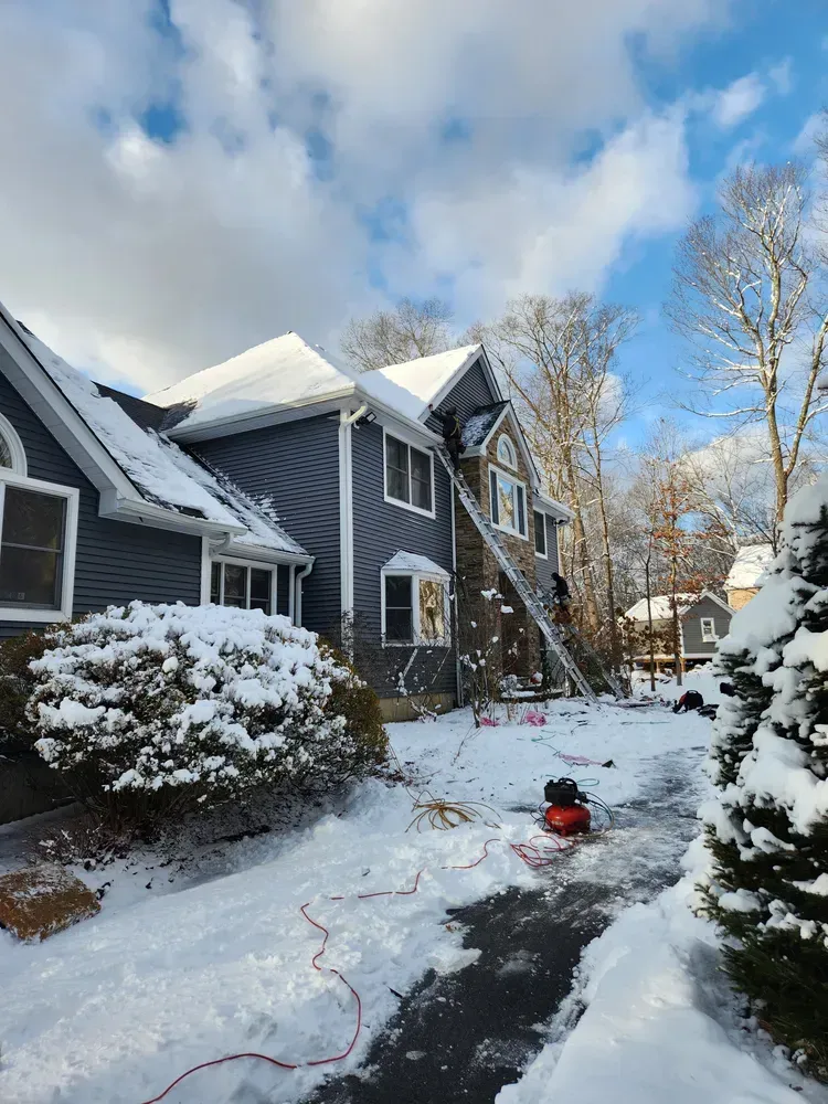 House with snow-covered roof and surroundings; ladder leaning against the side; winter scene.