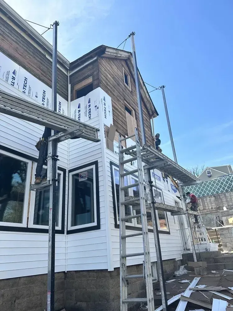 Construction workers installing siding on a two-story house, using scaffolding and ladders. Bright blue sky.