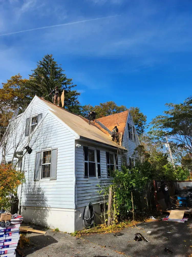 House under construction; workers on the roof removing old shingles. Blue siding, clear sky, and foliage surrounding.