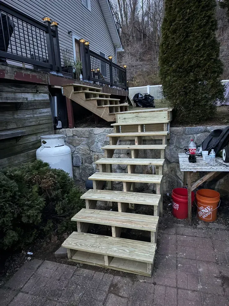 Wooden outdoor staircase leading up to a deck, set against a hillside and a house.