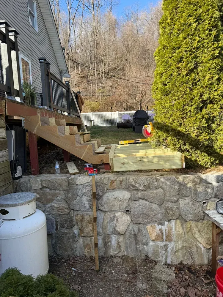 Wooden deck stairs under construction, set against a stone retaining wall in a yard.