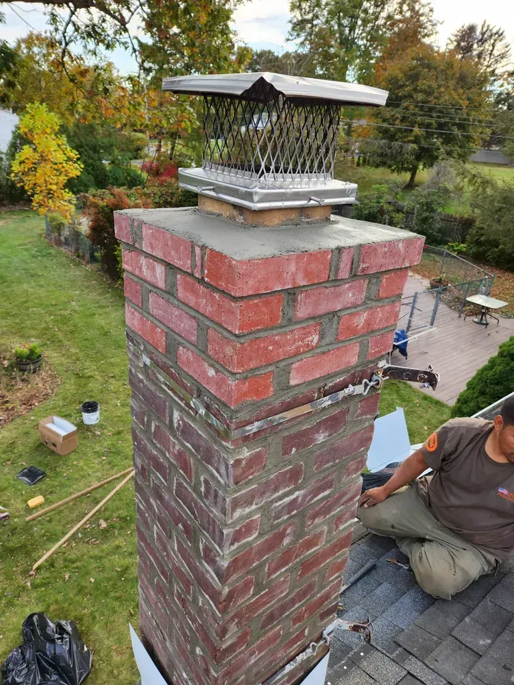 Chimney with a cap on a roof, being worked on by a person in a brown shirt.