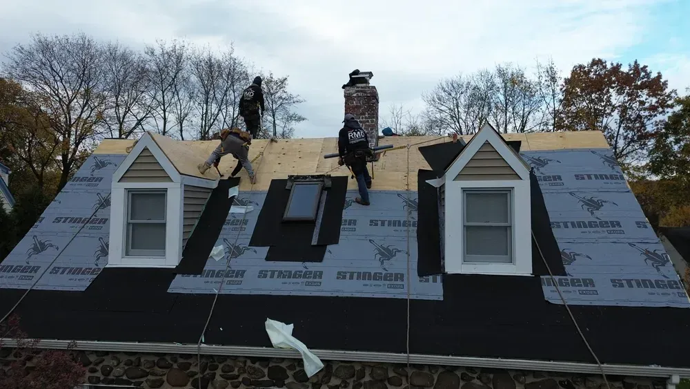 Roofers working on a house roof, laying down underlayment and shingles. Cloudy sky, trees in background.