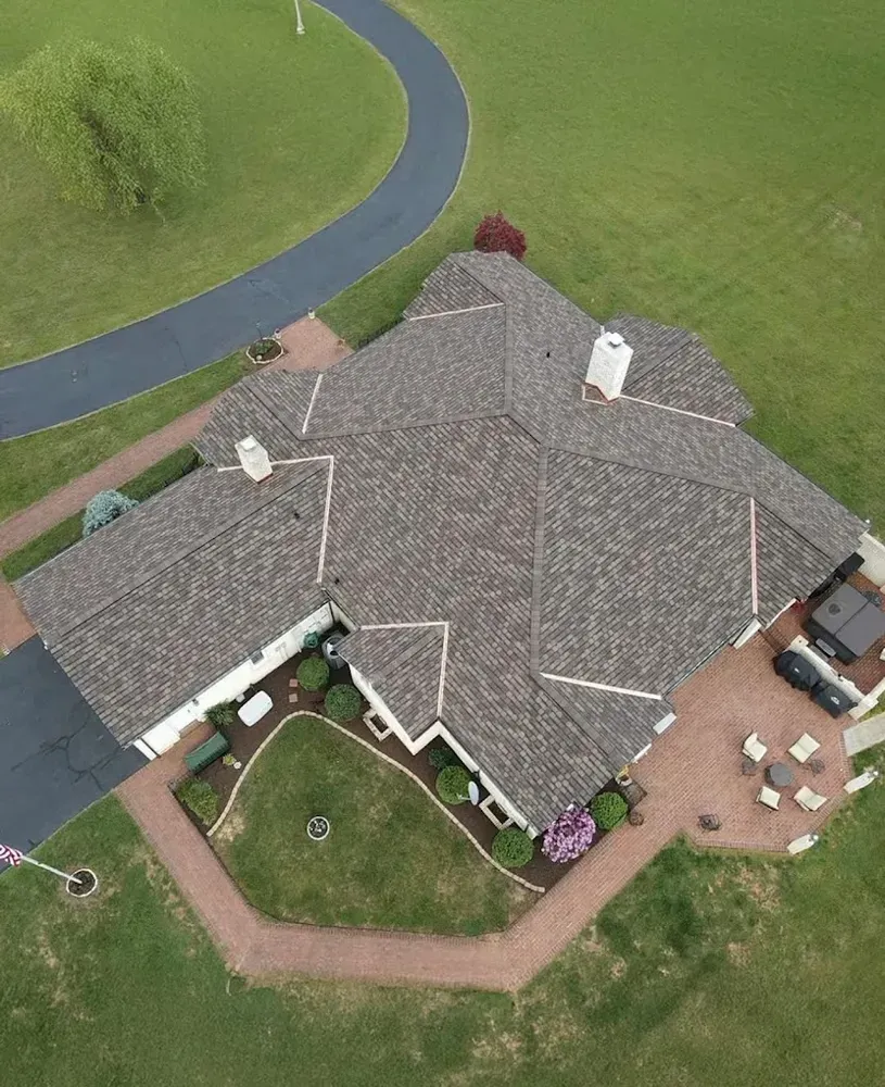 Aerial view of a large house with a complex roof, driveway, patio, and surrounding green lawn.