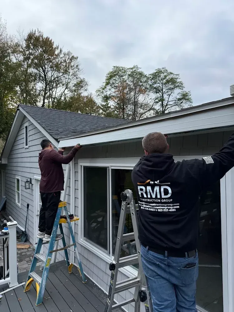 Two workers on ladders installing siding on a house with gray siding and dark roof. Cloudy sky.