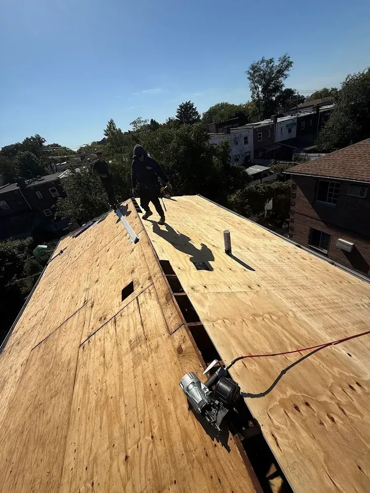 Workers on a rooftop, preparing for new roofing. Plywood and a nail gun are visible. Sunny day.