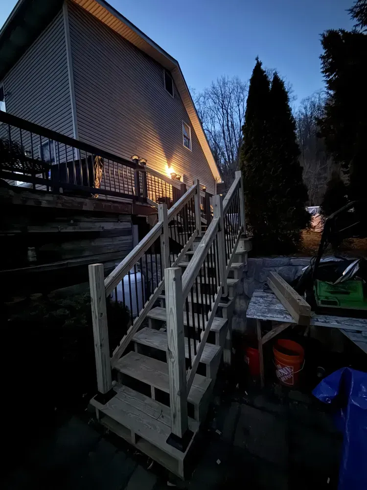 Outdoor wooden staircase leading up to a house at dusk. Black railings, gray steps, and a lighted deck above.