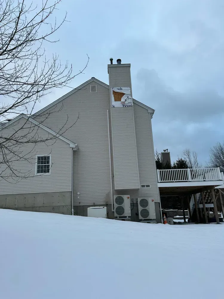 Snow-covered house with a chimney displaying a Wisconsin-shaped flag, flanked by HVAC units, under a cloudy sky.