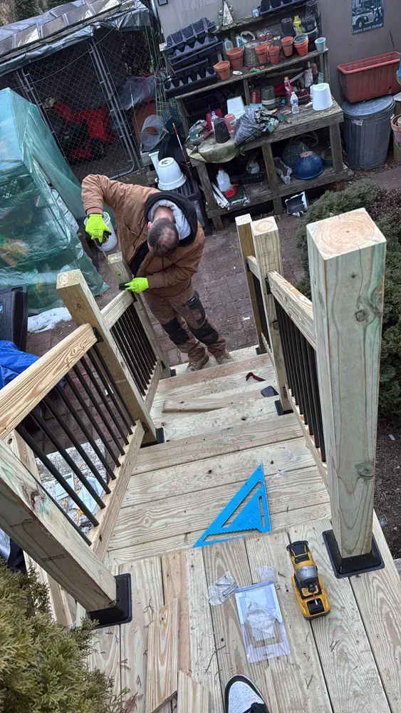 Man working on wooden stairs with black railings; tools on steps. Outdoor setting.