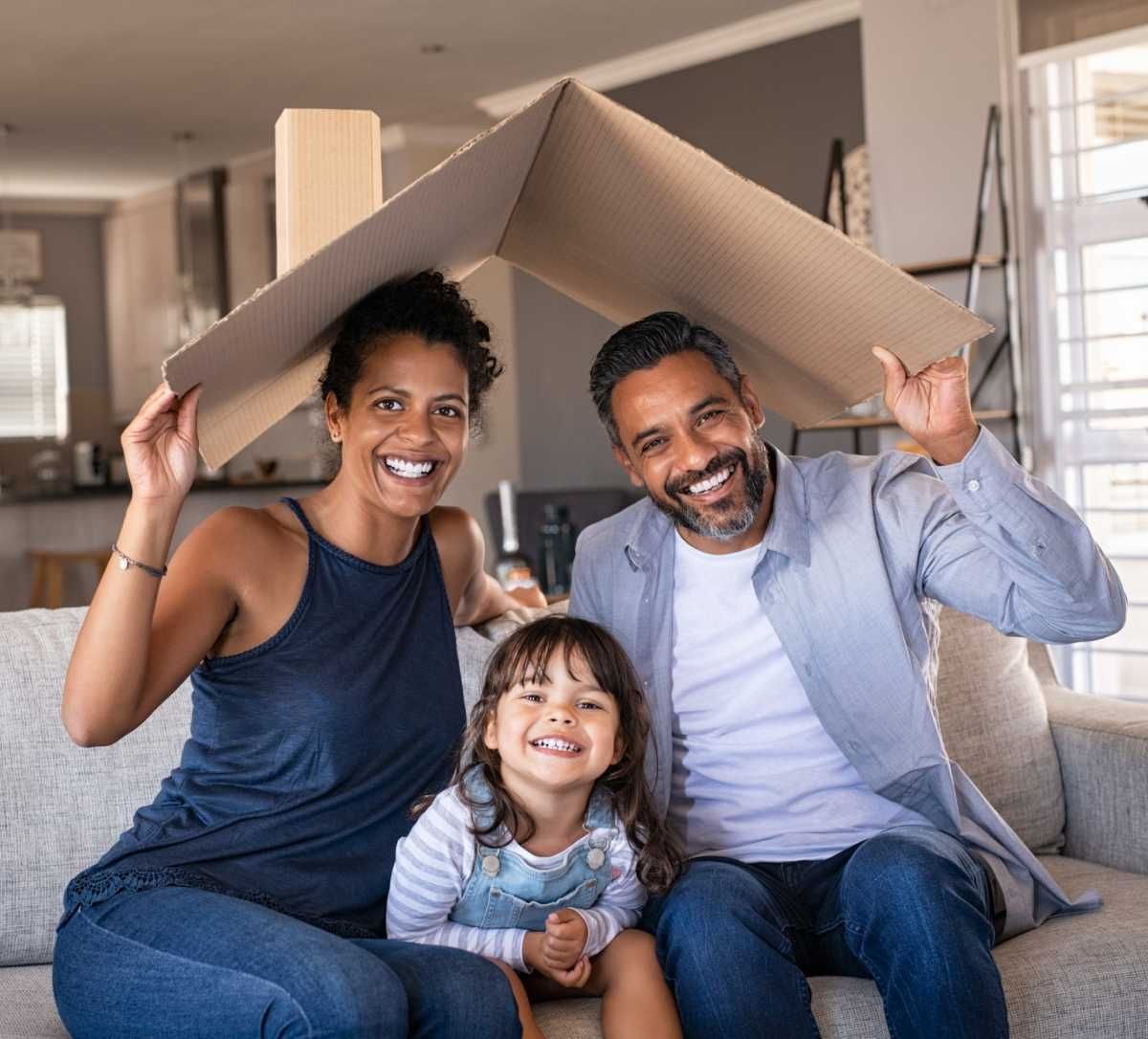 Family of three smiling, holding cardboard roof indoors.