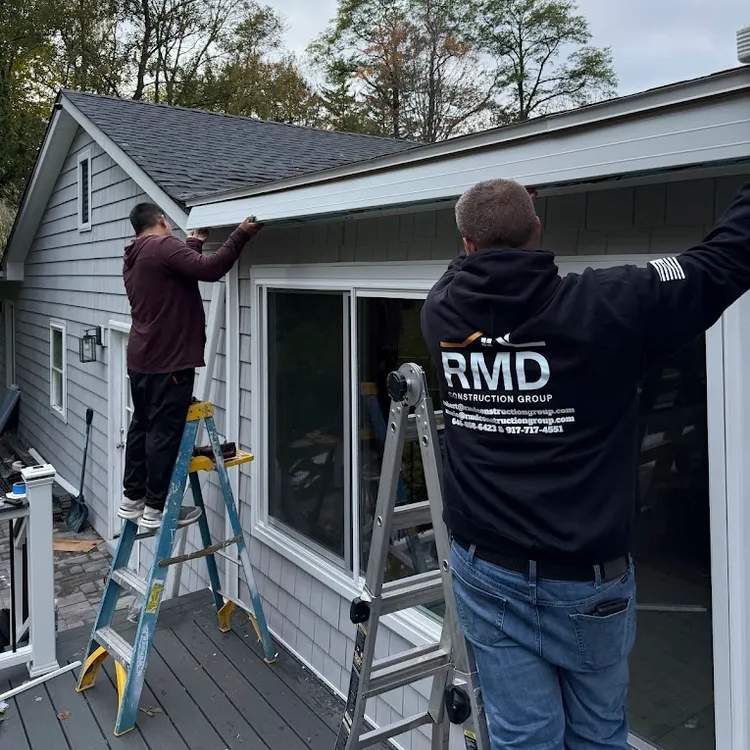 Two workers install gutter along the roof of a house with light grey siding. One is on a ladder.