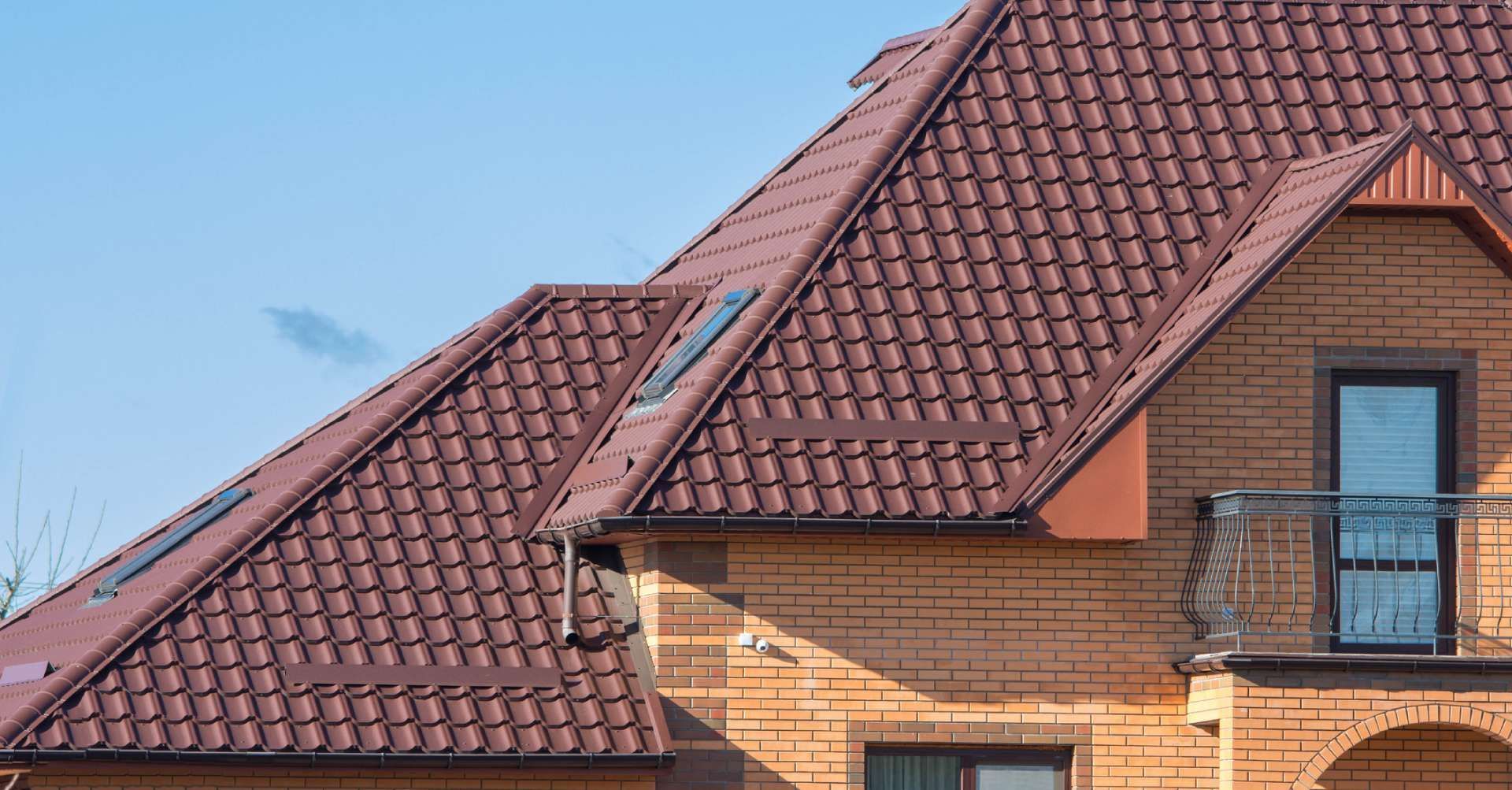 Brown roof on a light brown brick house with a small balcony and a blue sky in the background.