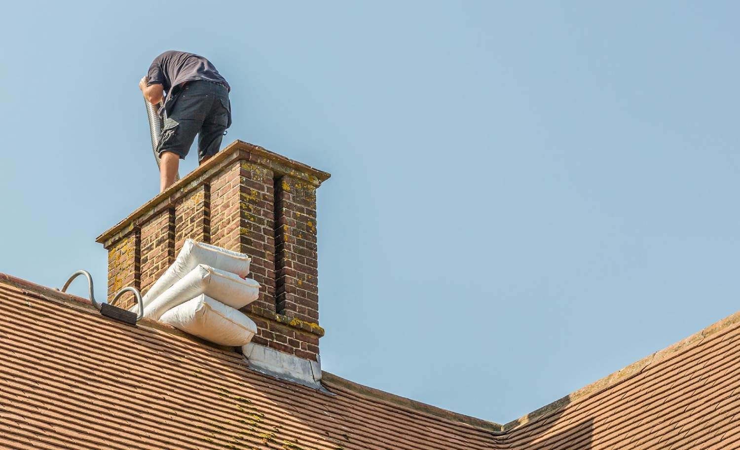 Person on a rooftop chimney, working. Blue sky. Brick chimney, terracotta roof.