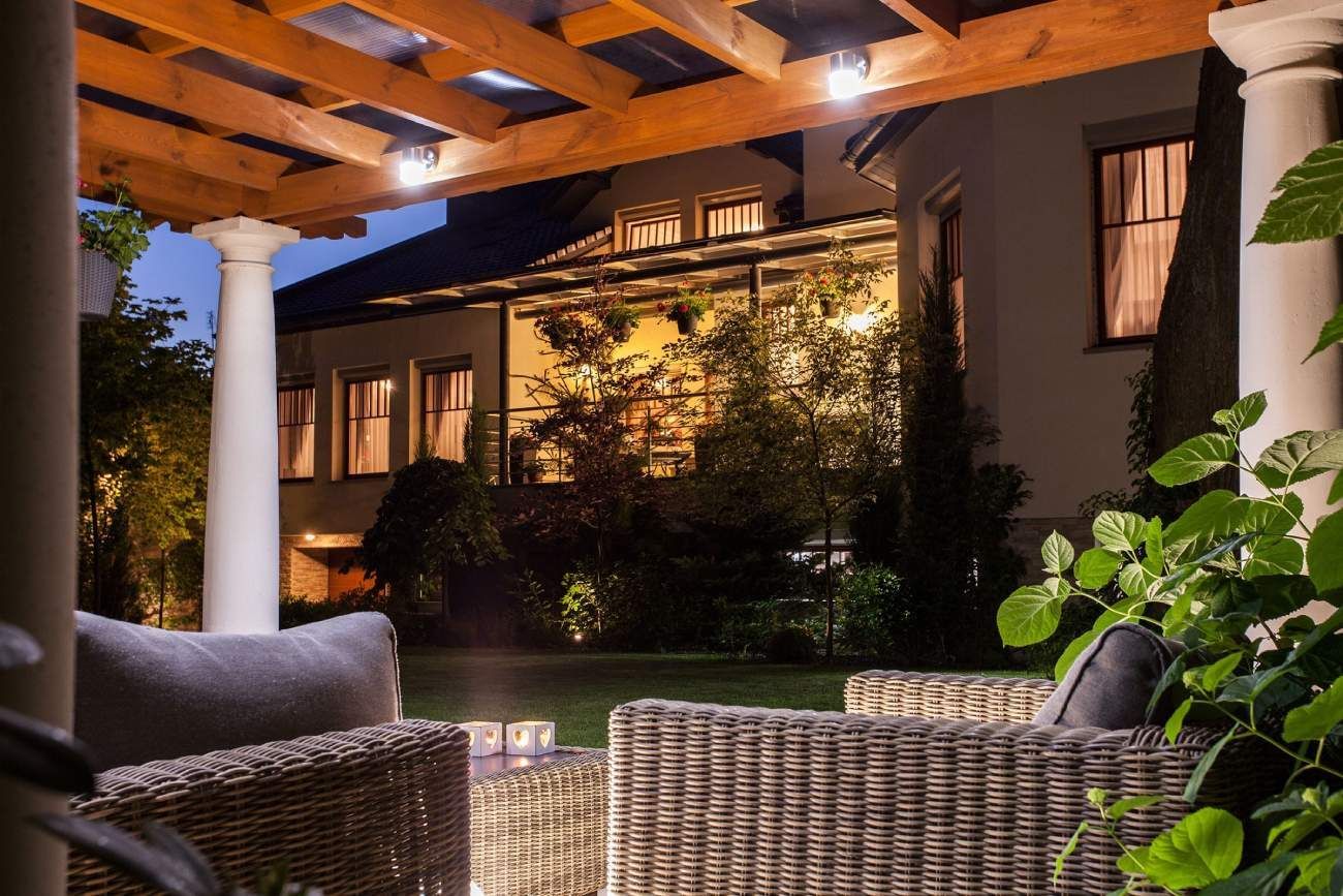 Outdoor patio with wicker furniture under wooden pergola, overlooking a house at dusk.