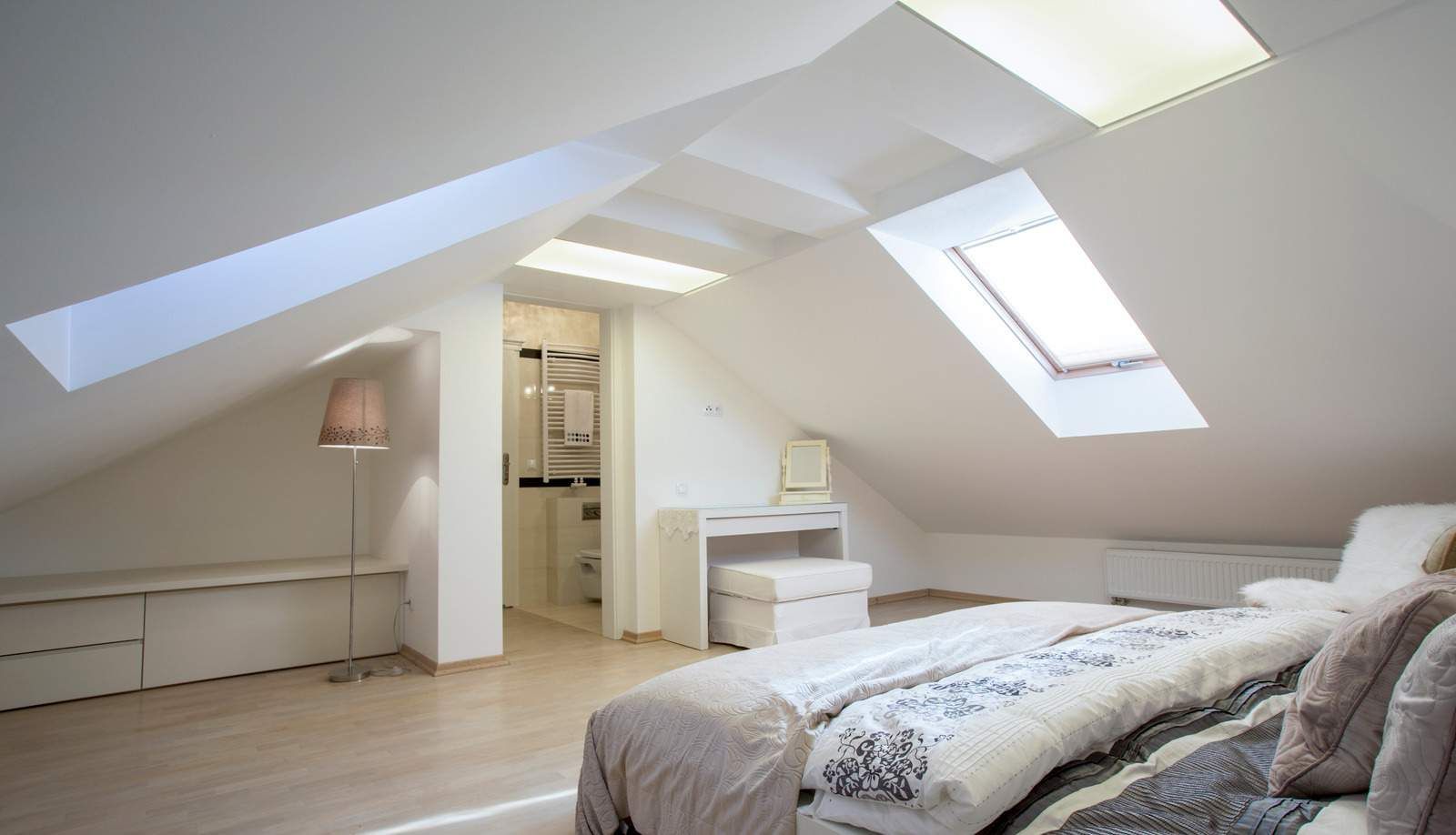 Bedroom in attic with skylights, white walls, bed with patterned cover, desk, bench, and doorway.