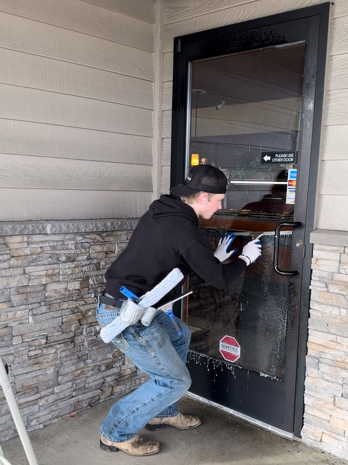 Man cleans a glass door with a squeegee; he's wearing a black hoodie, blue jeans, and gloves; outdoor setting.