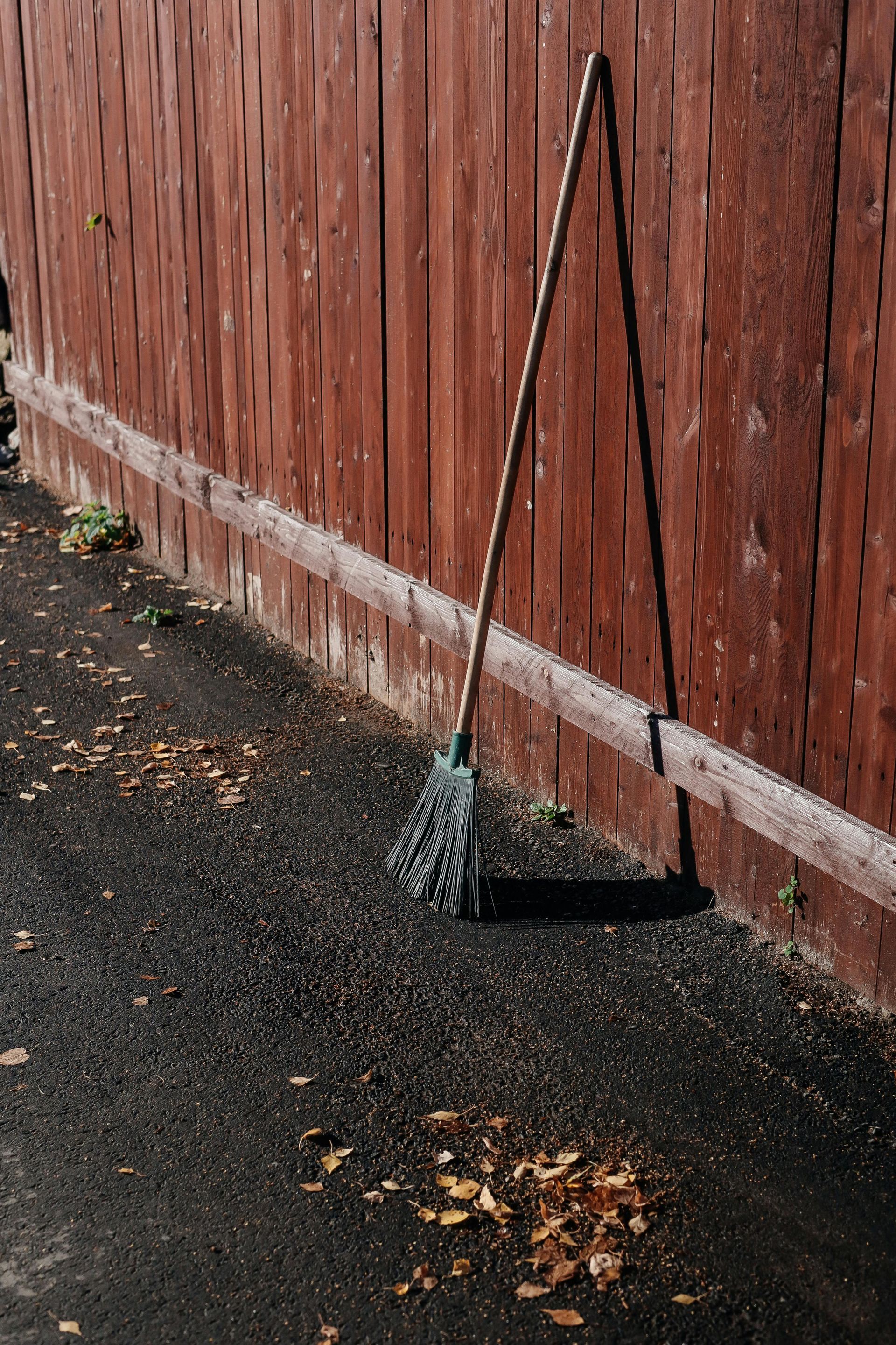 Mop leaning against a weathered brown wooden fence on an asphalt surface scattered with leaves.