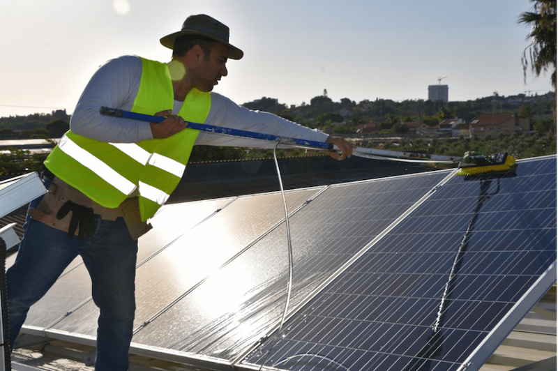 Man wearing safety vest cleans solar panels with a long brush on a rooftop.