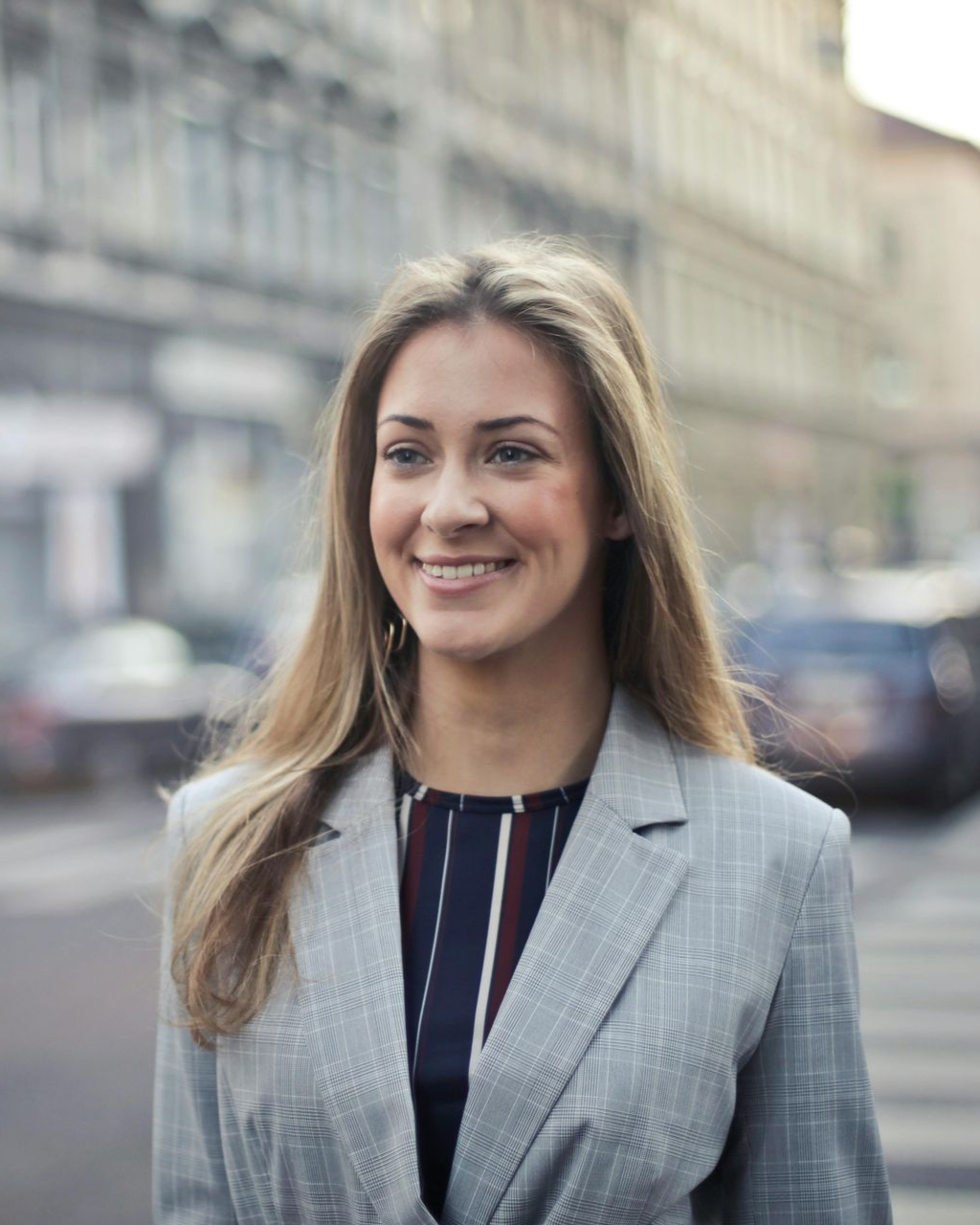 Woman in a gray blazer smiling, standing on a city street with cars and buildings in the background.