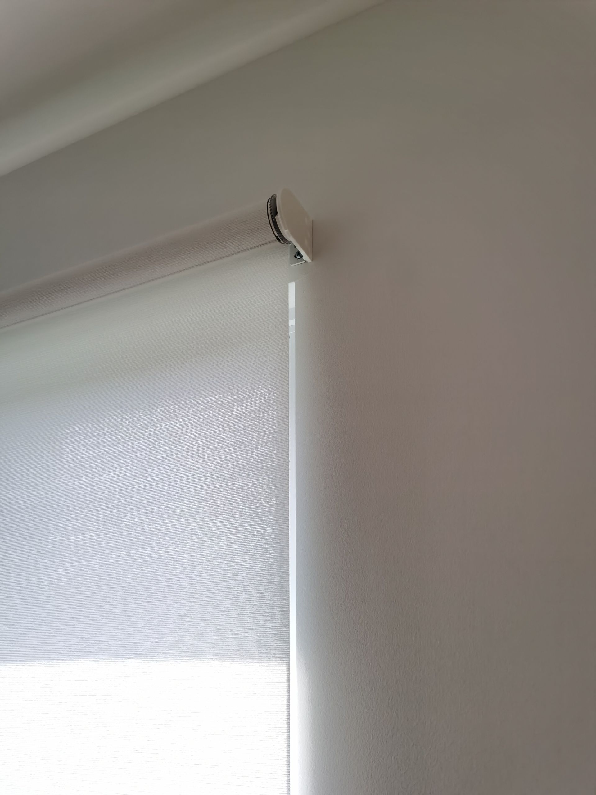 A white roller blind is hanging on a white wall next to a window.