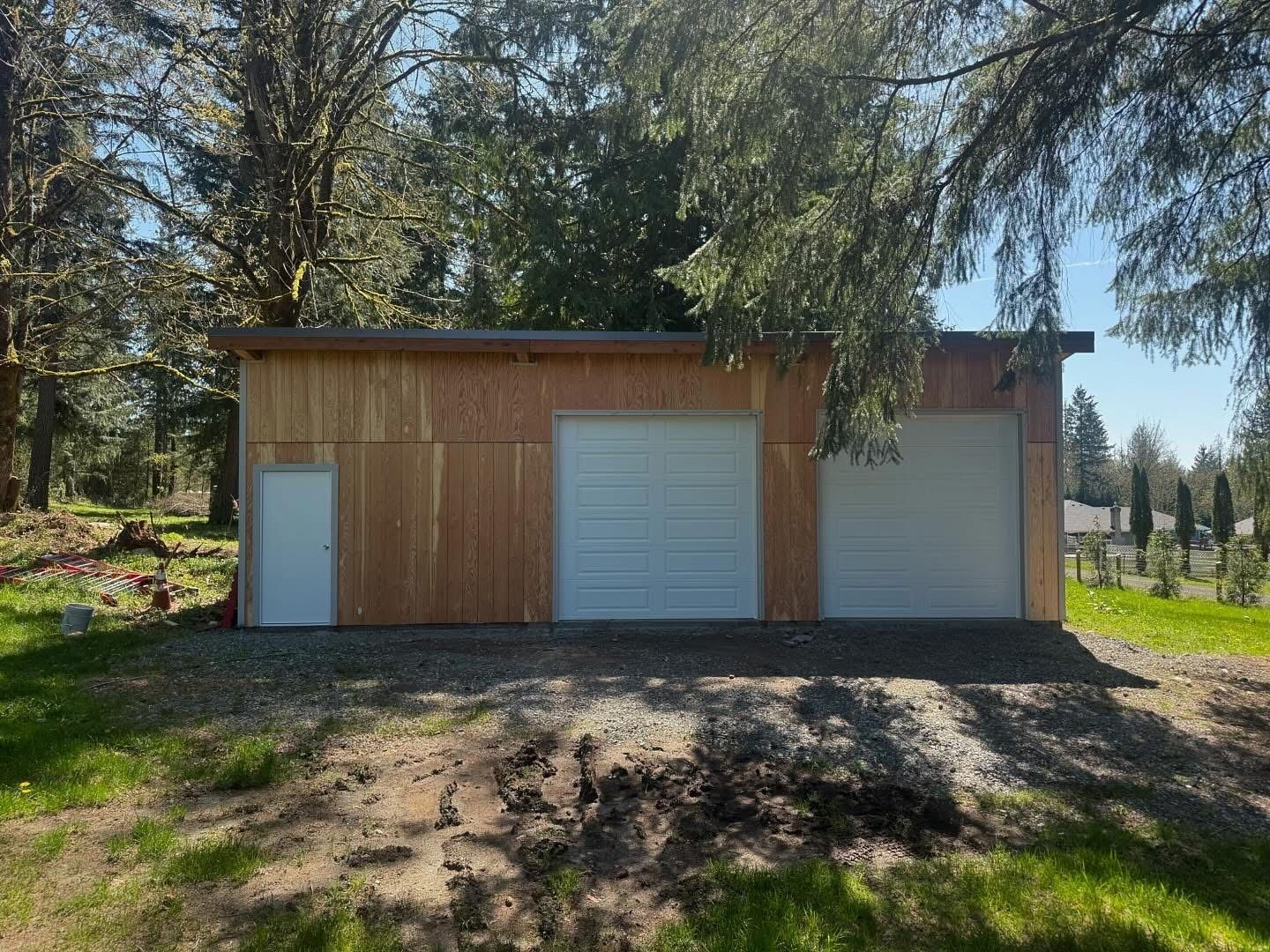A tan shed with white garage doors and a side door sits on a gravel bed in a grassy yard.