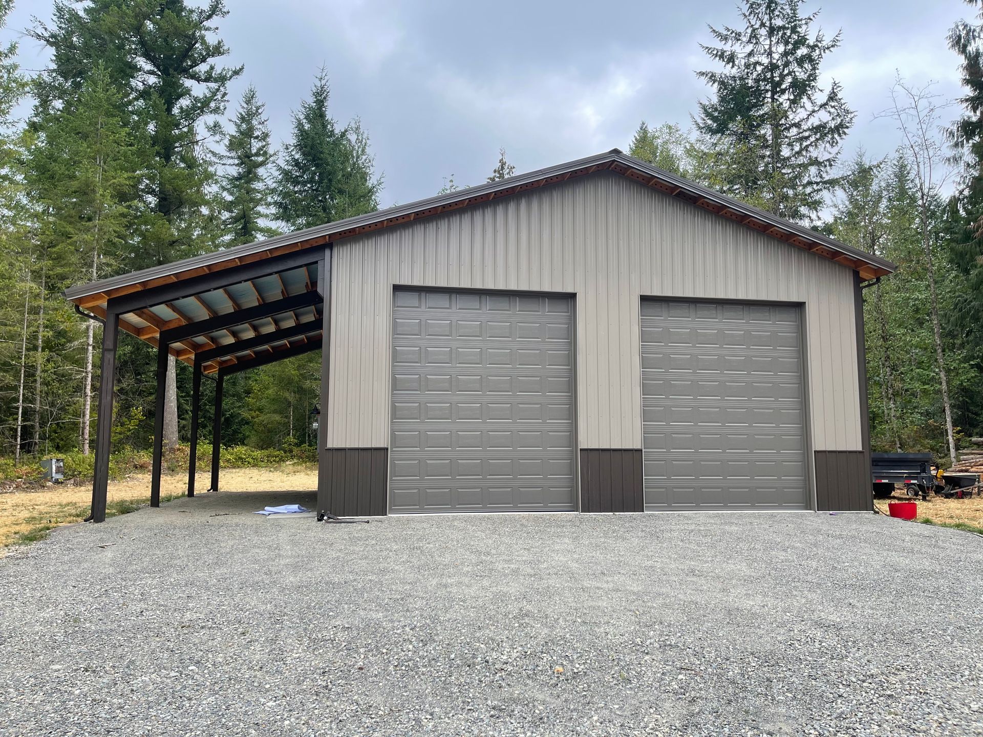Two-car metal garage with attached carport. Tan and brown exterior on a gravel lot, forested background.