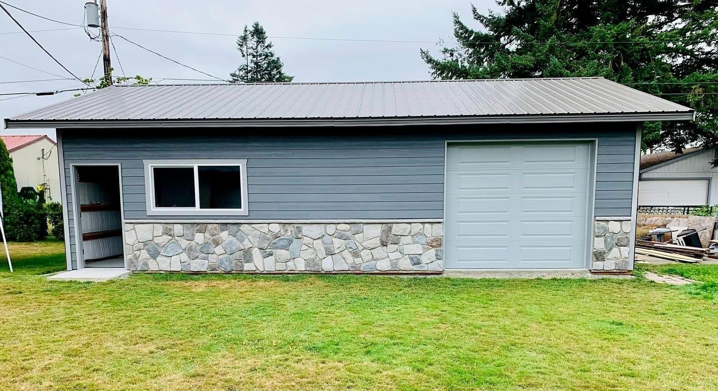 Garage with gray siding, stone facade, metal roof, and open storage on a lawn.