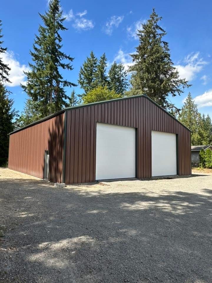 Brown metal garage with two white overhead doors on a gravel driveway, surrounded by trees.
