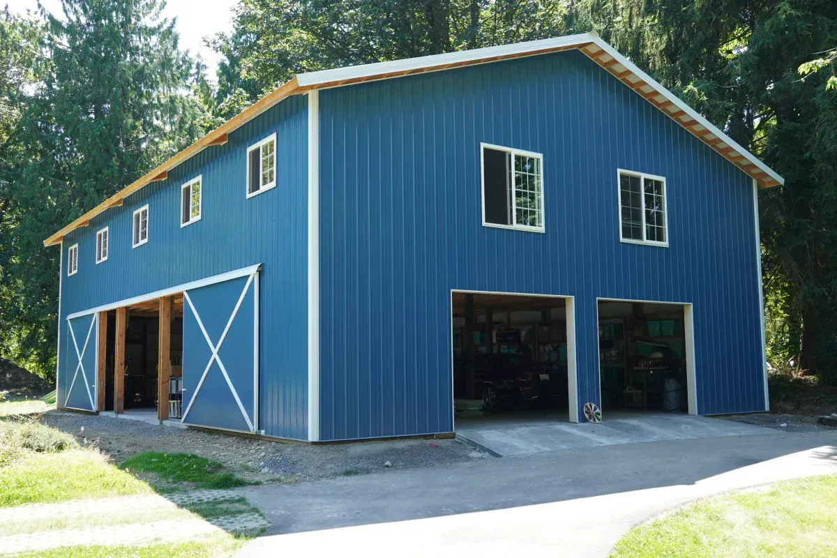 Blue barn with two garage doors, multiple windows, and sliding doors. Set in a wooded area.