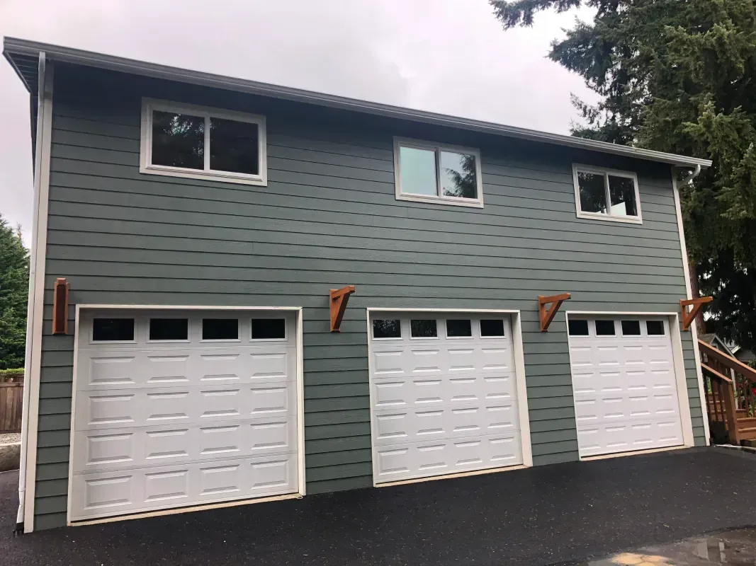Three-car garage with white doors, blue-green siding, and three upper windows on a cloudy day.